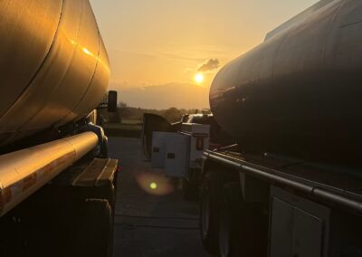 A wide-angle sunset view captures two large fuel tankers parked side-by-side, with their curved metallic surfaces reflecting the warm evening light. A service vehicle with open utility doors is positioned between the trailers, silhouetted against the glowing sun on the horizon.