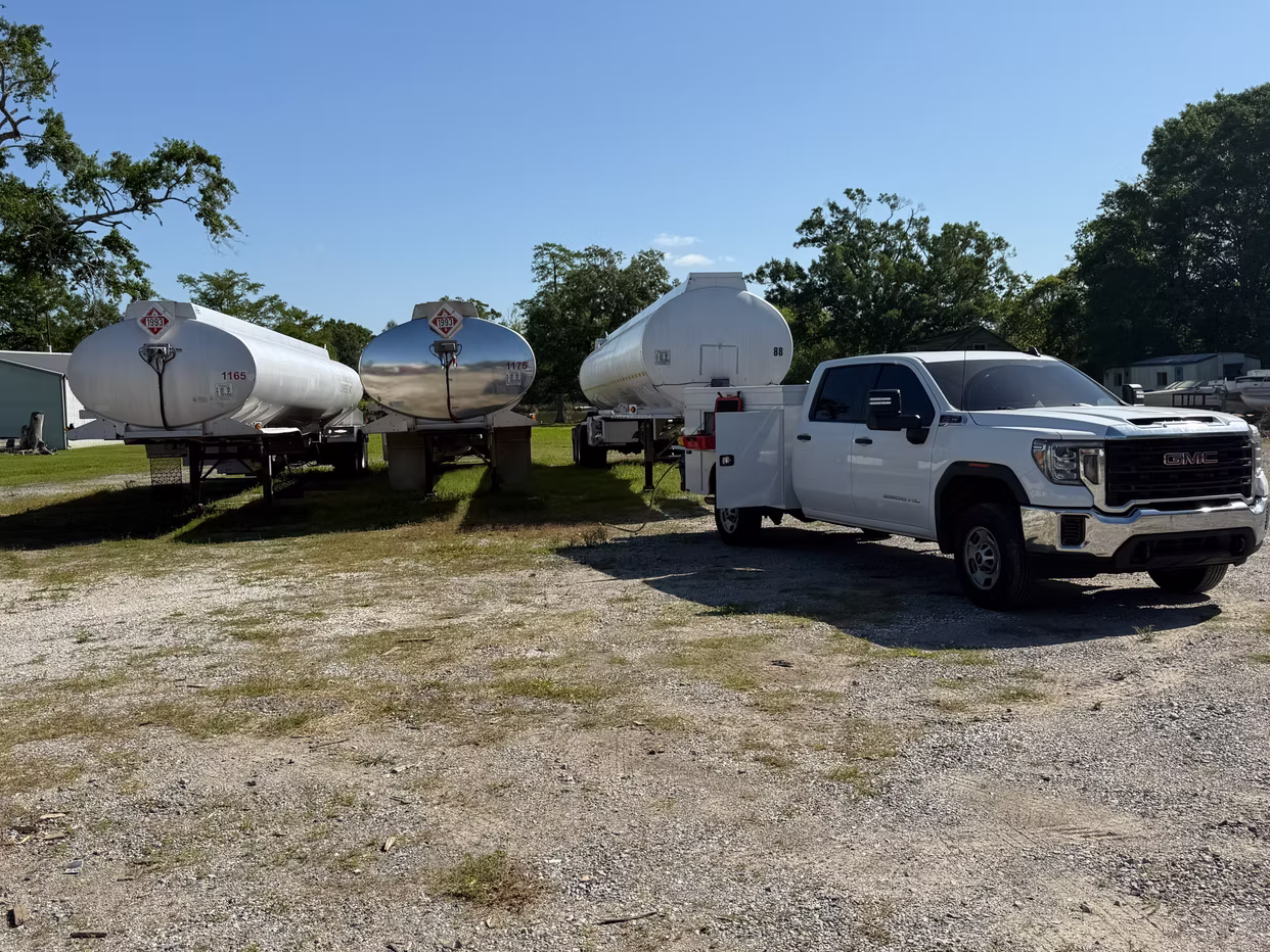 A white GMC service truck is parked in a gravel lot next to three large, white fuel tanker trailers under a clear blue sky. The tankers are positioned in a row, with the service truck's utility bed open as if prepared for maintenance or inspection tasks.