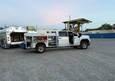 A white GMC service truck with open utility compartments is parked on a gravel lot next to the rear of a fuel tanker. The truck's doors and side cabinets are open, revealing tools and equipment being used for maintenance or inspection in an industrial setting.