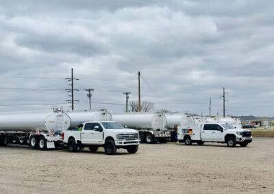 Two white service trucks, including a Ford and a GMC, are parked in a gravel lot alongside a row of large white fuel tanker trailers under an overcast sky. The trucks appear to be part of a mobile inspection or maintenance crew positioned for field service on the industrial trailers.