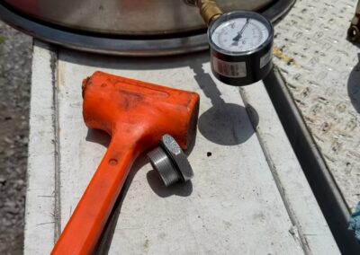 An orange dead-blow hammer and blue-handled pliers rest on a metal workbench next to an industrial pressure gauge connected to a stainless steel tank. The setup is typical for maintenance or testing of cargo tank components in the field.