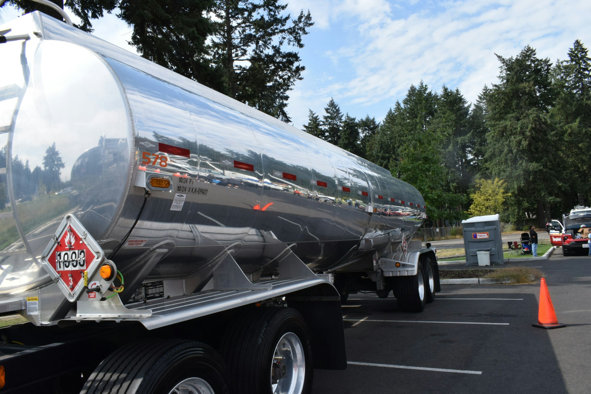 A shiny, polished silver fuel tanker trailer is parked in an asphalt lot with a dense line of evergreen trees in the background. The rear of the trailer displays a red and white hazardous materials placard with the number "1993" and a small orange safety cone is positioned on the ground nearby.