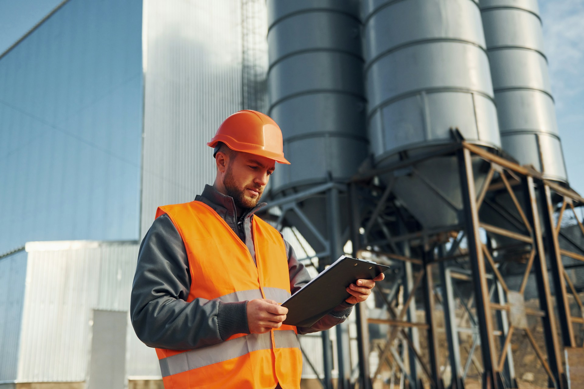 An inspector wearing an orange hard hat and safety vest reviews notes on a clipboard in front of large industrial silos. The technician is positioned outdoors at a facility, likely conducting a site safety audit or equipment inspection.