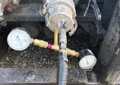 A close-up view of an industrial setup shows two pressure gauges connected to a central valve assembly via a brass T-junction. A yellow hose extends from the bottom of the assembly, while the gauges provide readings for the system which is attached to a large, bolted metal pipe.