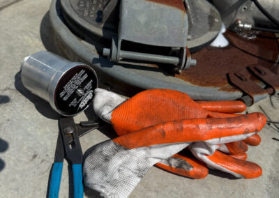 A pair of blue-handled pliers and orange work gloves sit on the metal surface of a tanker truck next to a rusted access hatch. A small cylindrical Betts component lies nearby, suggesting ongoing maintenance or inspection of the tank's hardware.