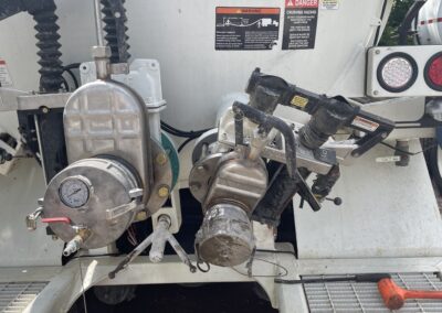 The rear of a white service vehicle features complex industrial pumping equipment, including a primary valve housing with an integrated pressure gauge and various lever controls. Warning labels regarding crushing hazards and vacuum safety are visible on the tank's surface, positioned above a metal grate walkway.