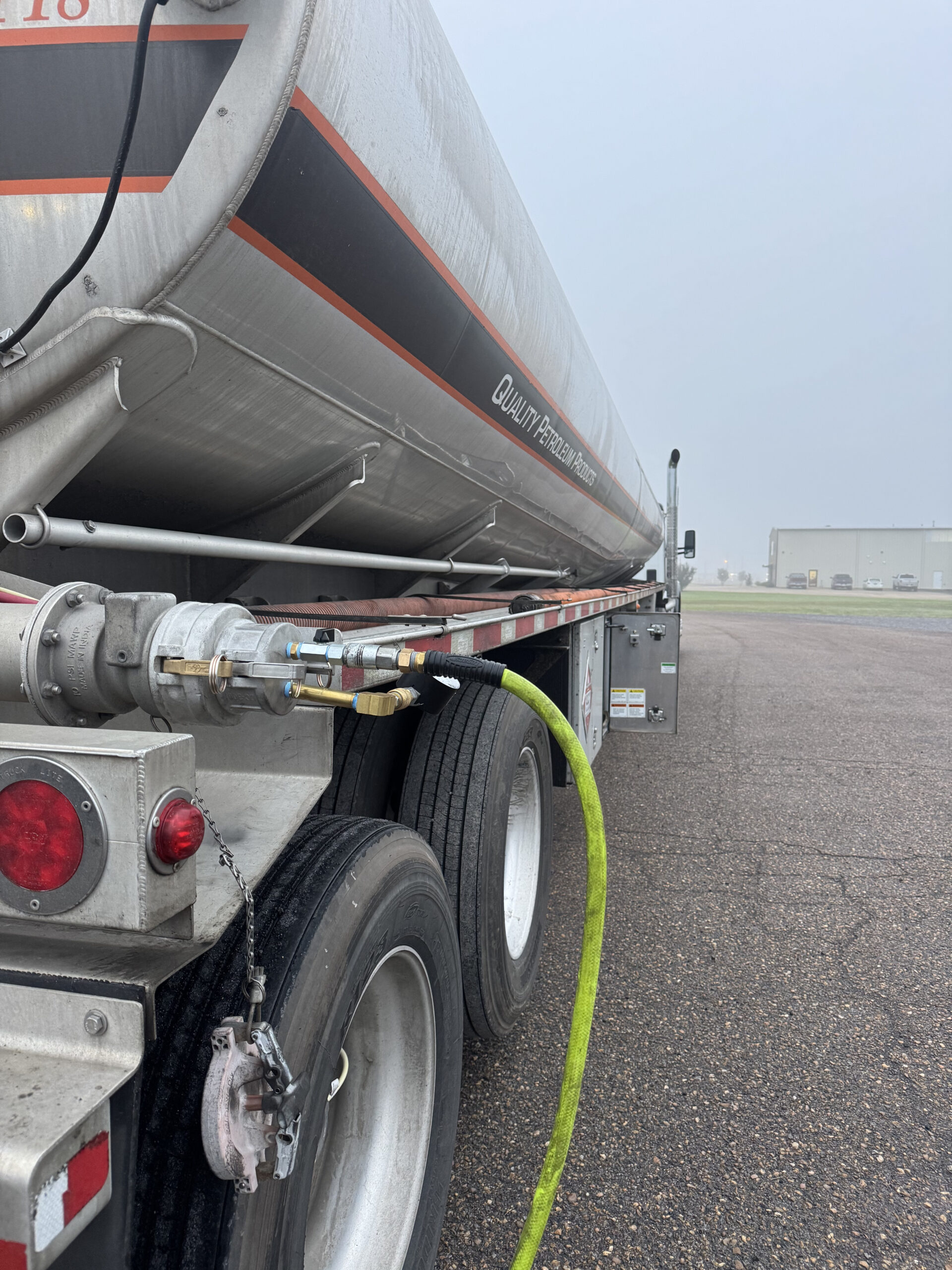 A side view of the tanker truck shows a bright green hose connected to a silver metal valve assembly on the rear chassis. The trailer features a black and orange stripe with the words "QUALITY PETROLEUM PRODUCTS" printed along the side.