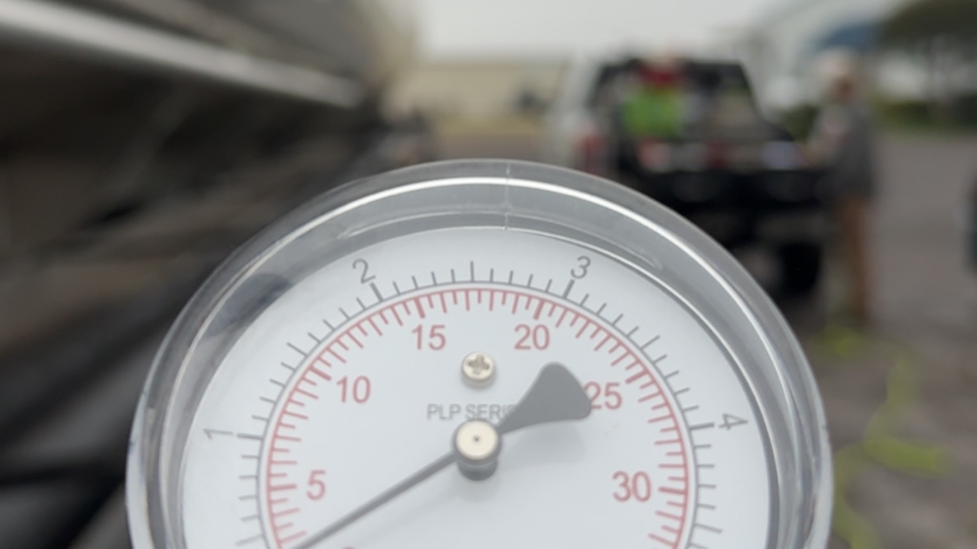 A close-up shot features a circular pressure gauge with a needle pointing to approximately 24 on a red scale. The background is blurred, showing a service vehicle and workers in a field, suggesting a technical or industrial inspection in progress.