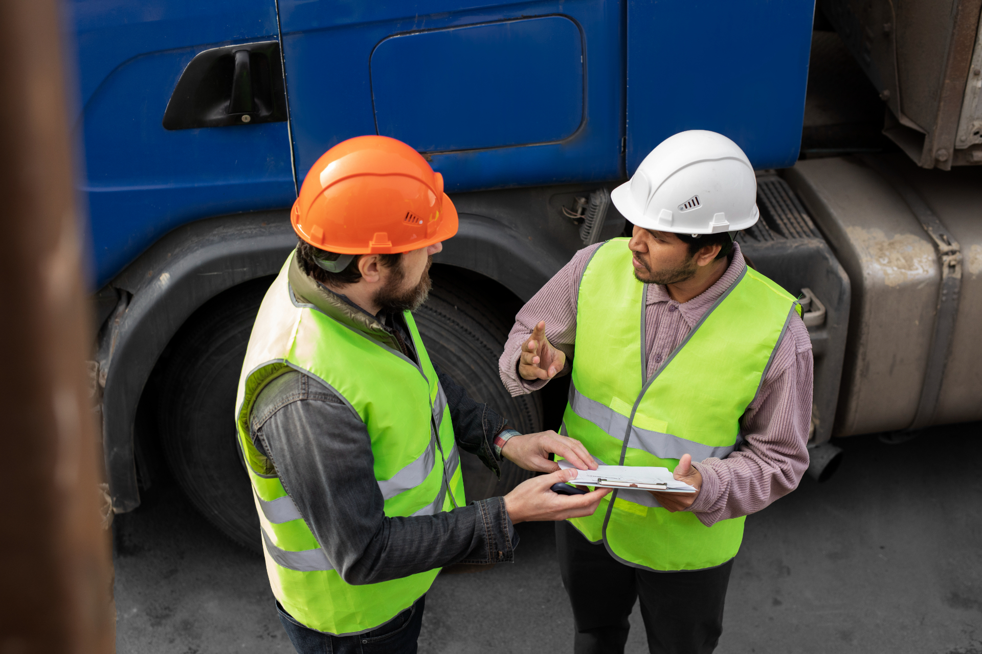 Two tank inspectors wearing hard hats and high-visibility vests stand next to a large blue semi-truck while discussing paperwork on a clipboard. One man gestures with his hand as they review the documents together during a delivery or inspection.